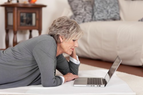 Жінка пенсіонер лежить на підлозі навпроти ноутбука і шукає роботу через Інтернет