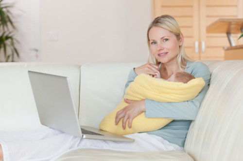 Молода мама сидить на дивані з ноутбуком на колінах і дитиною на руках